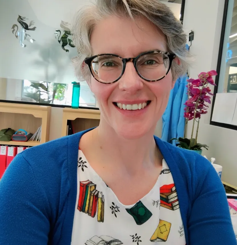 An image of Laura in her library office. She is a Caucasian woman wearing glasses with round tortoiseshell frames. She is wearing a white dress with books on it and a blue cardigan.